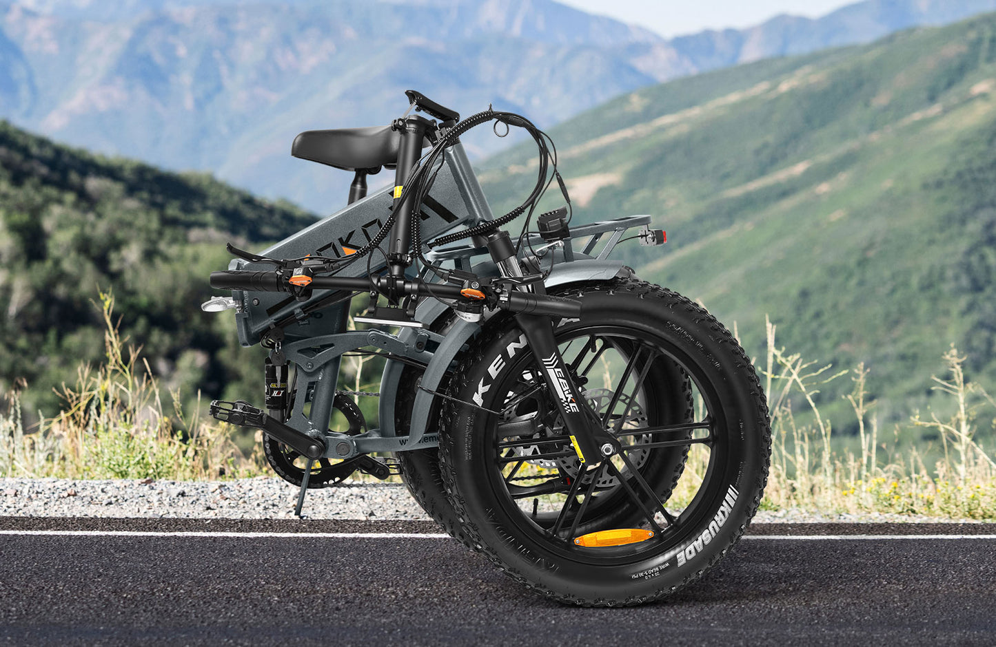 Folded gray electric bike with thick Kenda tires on asphalt road with green mountains background