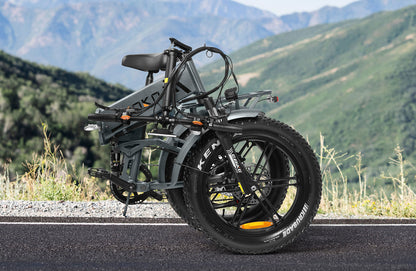 Folded gray electric bike with thick Kenda tires on asphalt road with green mountains background